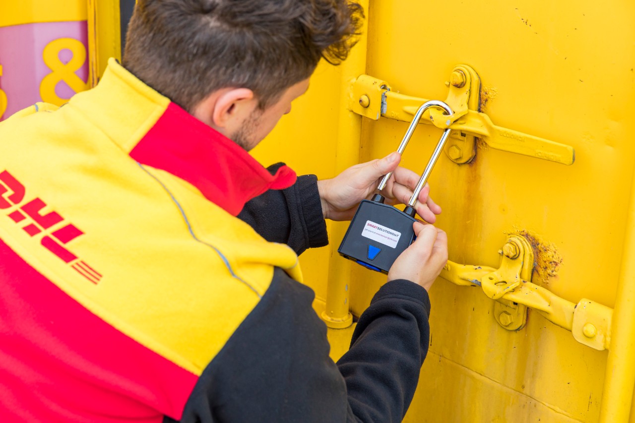 Männlicher DHL-Mitarbeiter in Uniform verschließt einen gelben Container mit der Hand. 